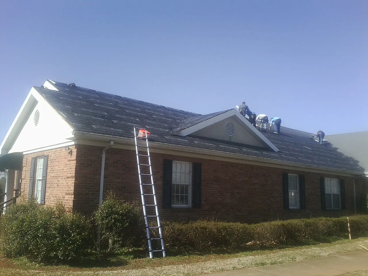 Crew tearing shingles off a brick ranch roof with a ladder set — Metal Roof Repair work by our crew in Colton